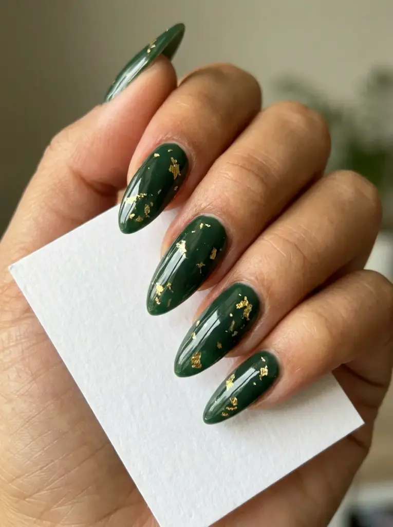 Glossy dark green almond manicure with gold foil flakes on long nails, close-up hand shot