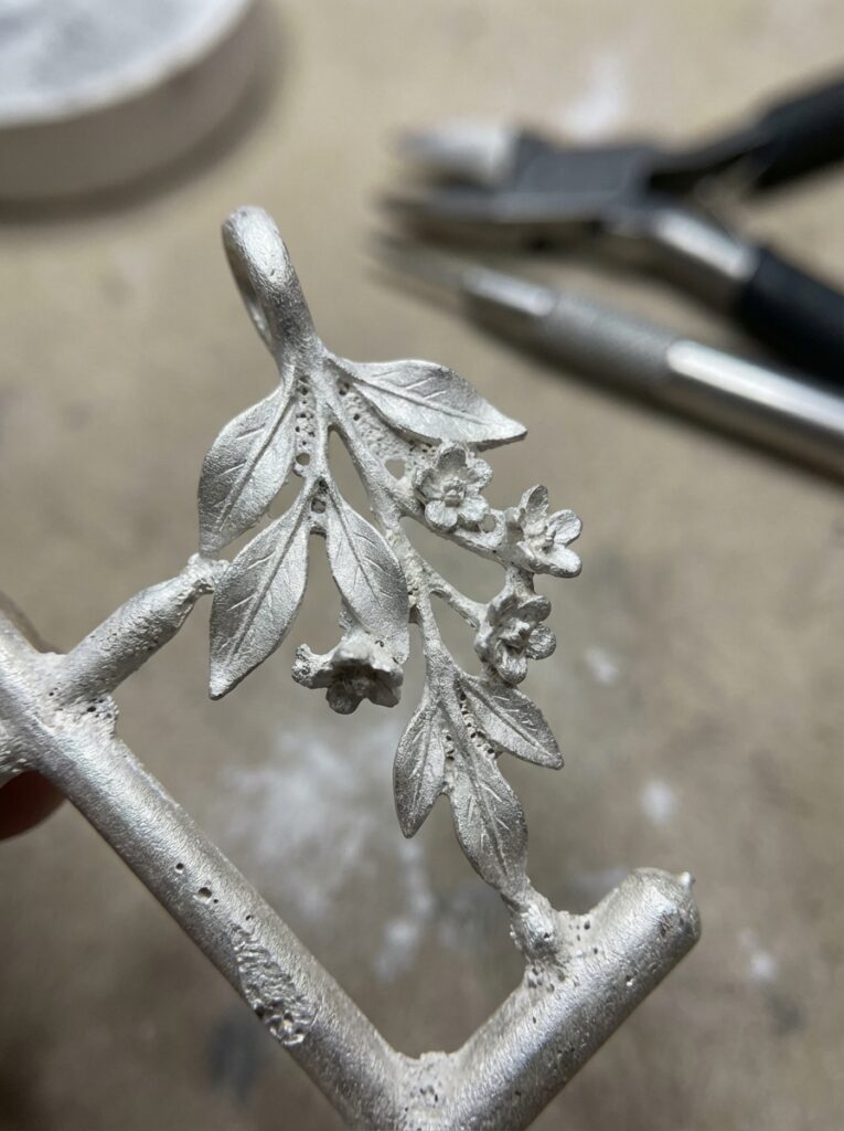 Close-up of handmade metal floral pendant casting for jewelry, detailed leaves and small blossoms on jeweler's bench