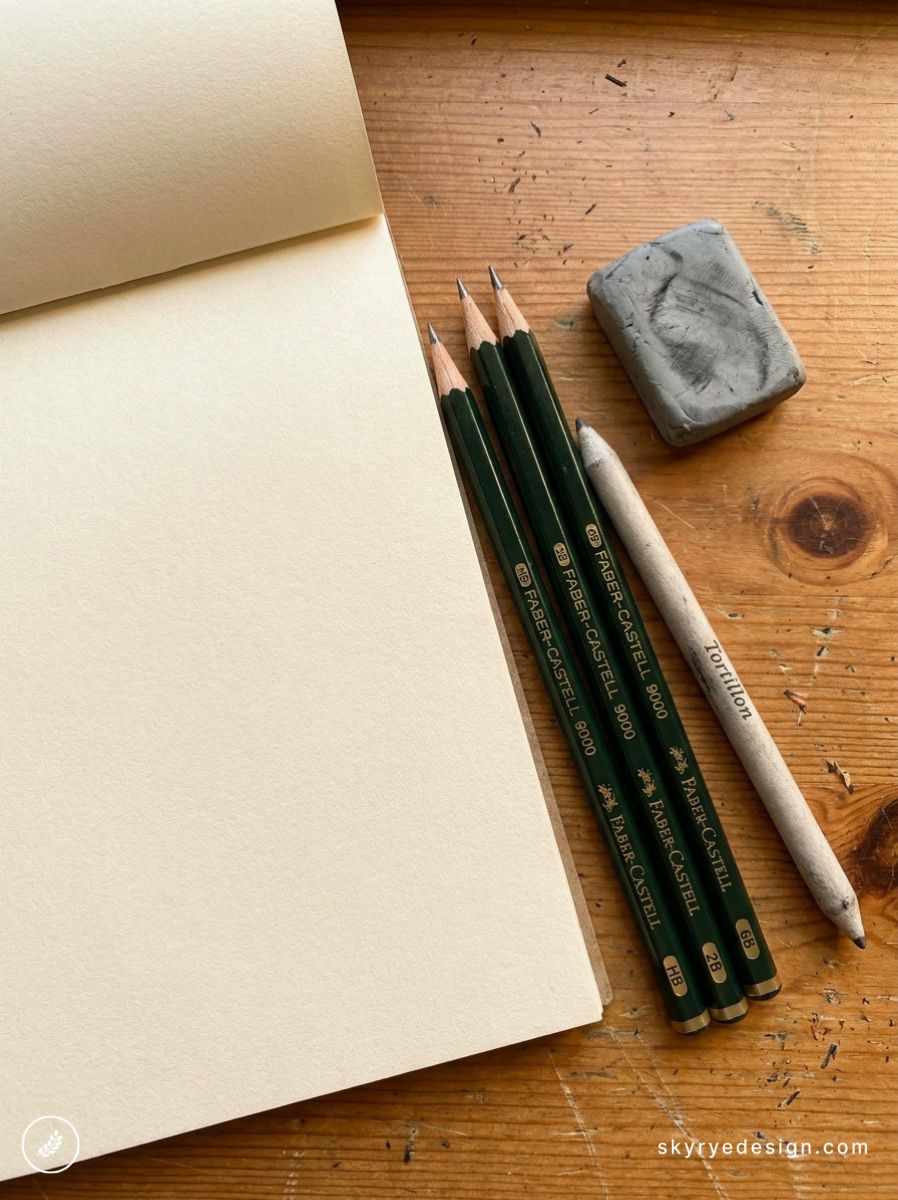 Blank sketchbook with Faber-Castell pencils, blending stump and kneaded eraser on wooden desk — drawing supplies.