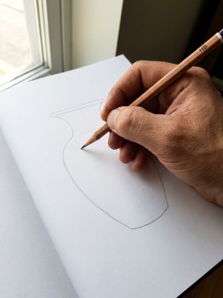 Hand sketching a vase outline with an HB pencil in a sketchbook by a sunlit window