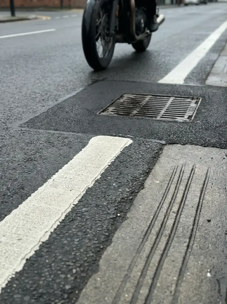 e3a4f027-f6b7-4cc7-bf98-dcb46f920e71 Wet asphalt close-up with white lane marking, storm drain grate, tactile paving grooves and motorcycle wheel | Sky Rye Design Wet asphalt close-up with white lane marking, storm drain grate, tactile paving grooves and motorcycle wheel