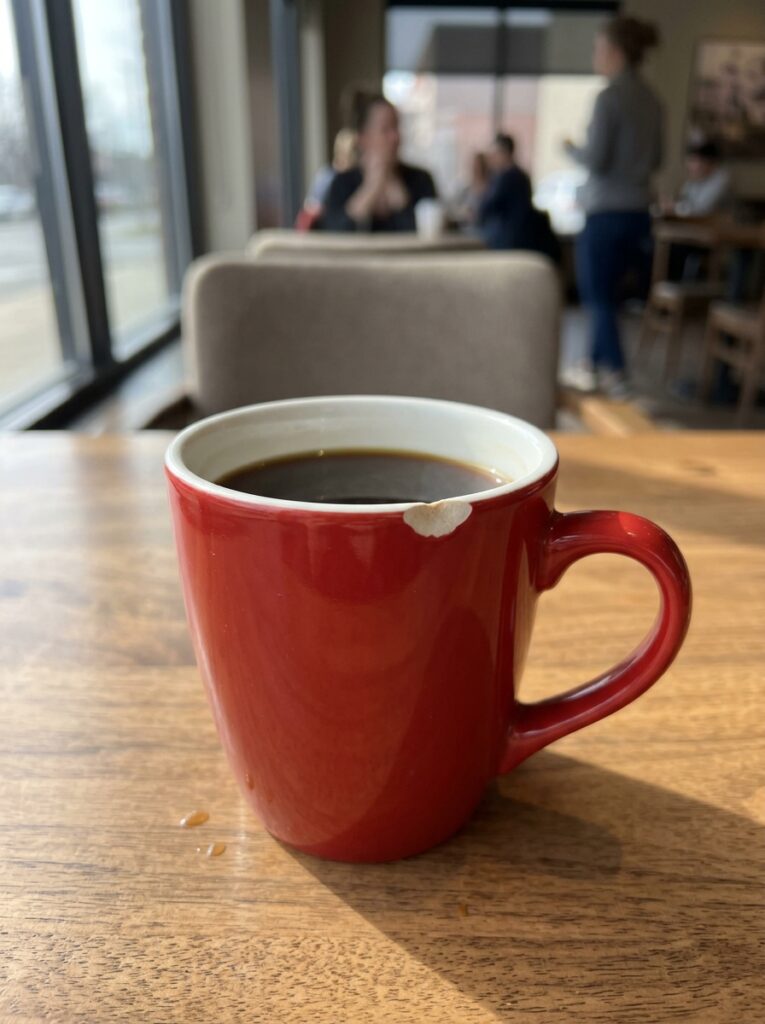 Red coffee mug with chipped rim filled with black coffee on wooden table in a busy cafe