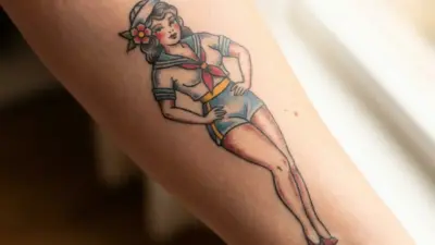 Colorful forearm tattoo of a vintage pin-up sailor girl in blue shorts, red neckerchief and flower hairpiece