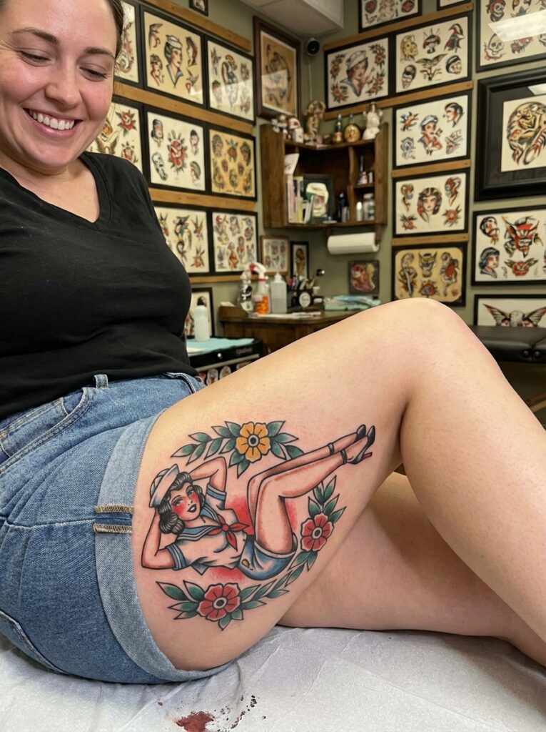 Smiling woman displaying fresh traditional American sailor pin-up thigh tattoo with floral wreath in tattoo studio