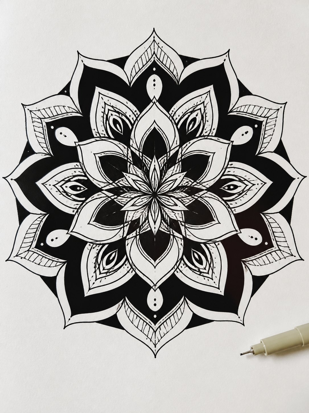 16 Mandala with Negative Space Intricate black-and-white mandala with layered petals and a drawing pen at the bottom right corner. | Sky Rye Design 16. Mandala with Negative Space mandala design on paper