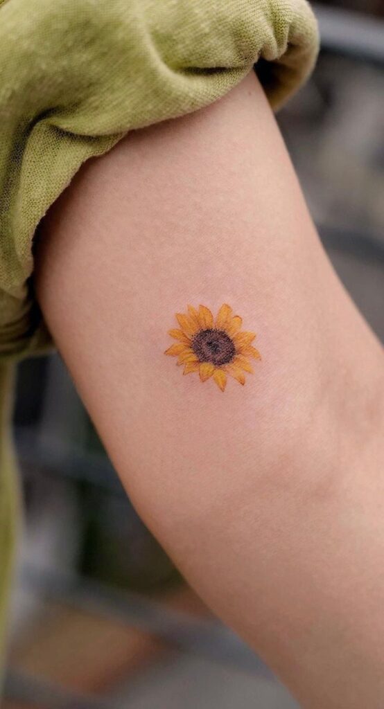 Tiny sunflower tattoo on inner arm, close-up showing yellow petals and brown center in minimalist ink