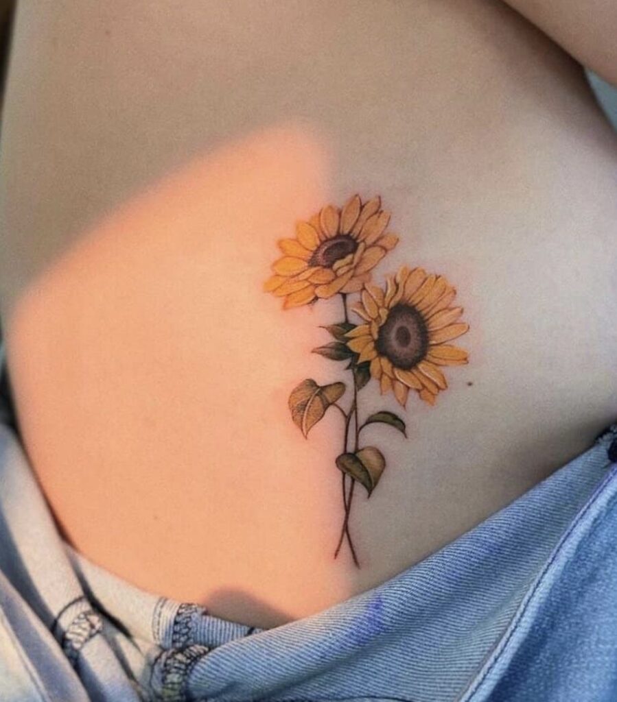 Sunflower tattoo: two yellow sunflowers on side rib above low-rise jeans, delicate stems and leaves