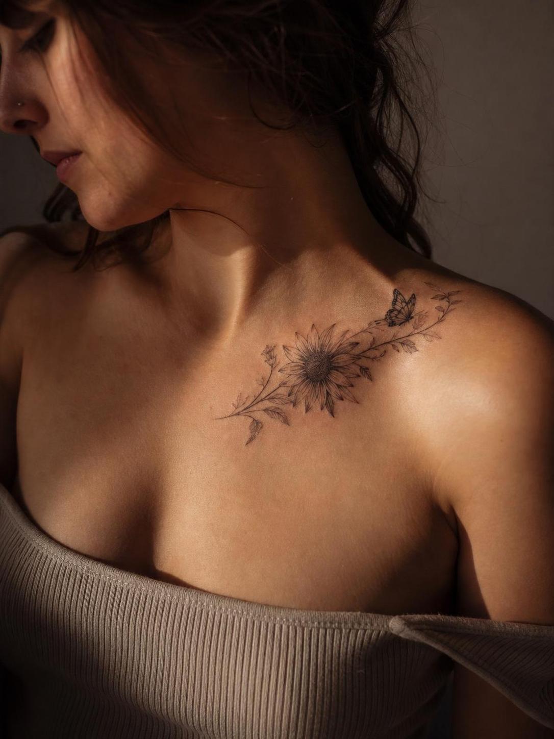 Sunflower collarbone tattoo on woman, fine-line butterfly and vines, close-up shoulder shot in natural light