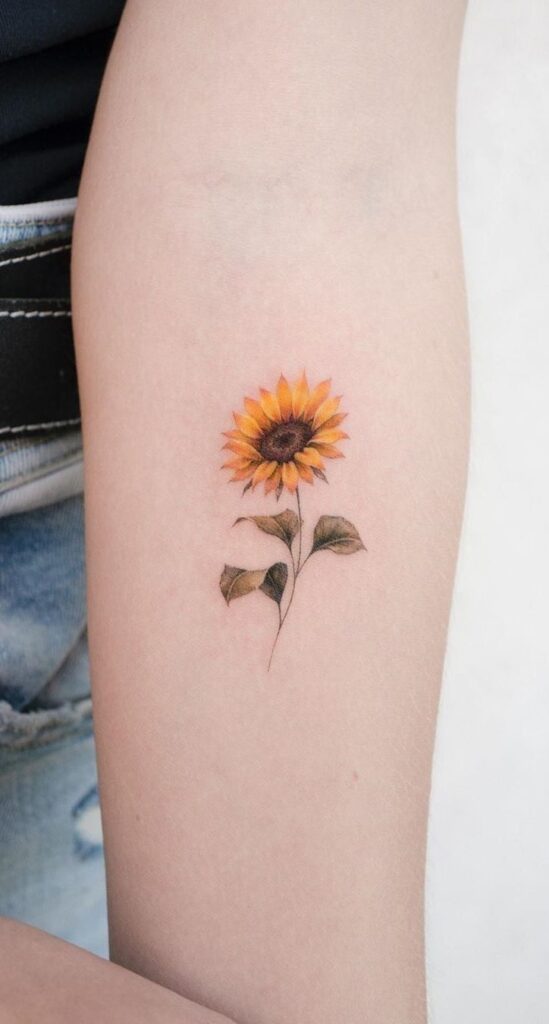 Small delicate sunflower tattoo with yellow petals and green leaves on inner forearm, fine-line minimalist floral ink