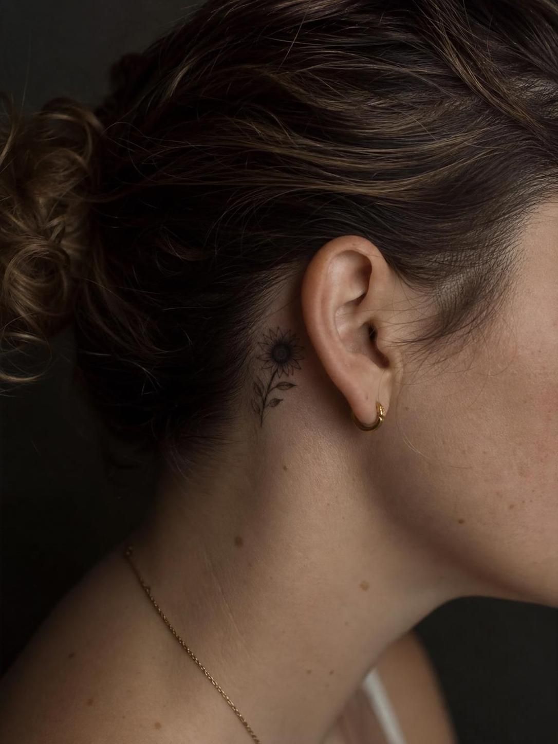Close-up profile showing small sunflower tattoo behind ear, gold hoop earring, and hair tied in a bun