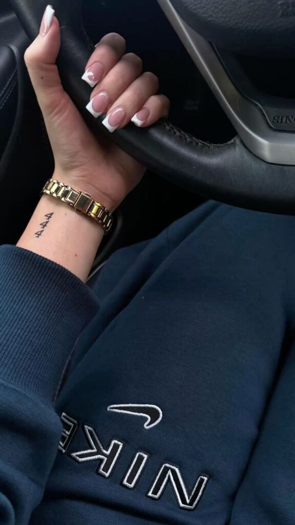 French manicure nails on hand holding car steering wheel, wrist '444' tattoo, gold watch, navy Nike sweatshirt