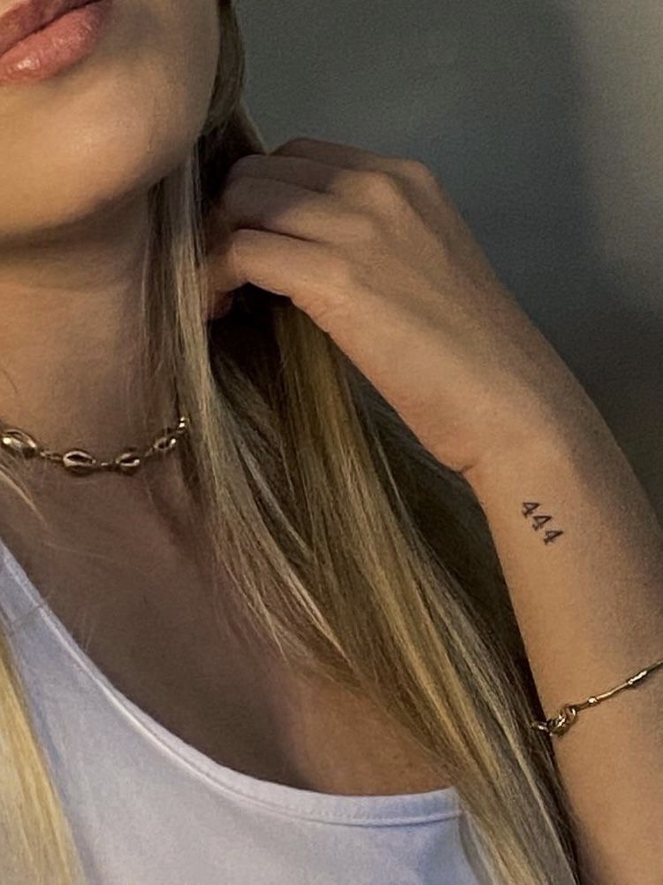 Close-up of woman with '444' wrist tattoo, gold shell necklace and bracelet, blonde hair, white tank top