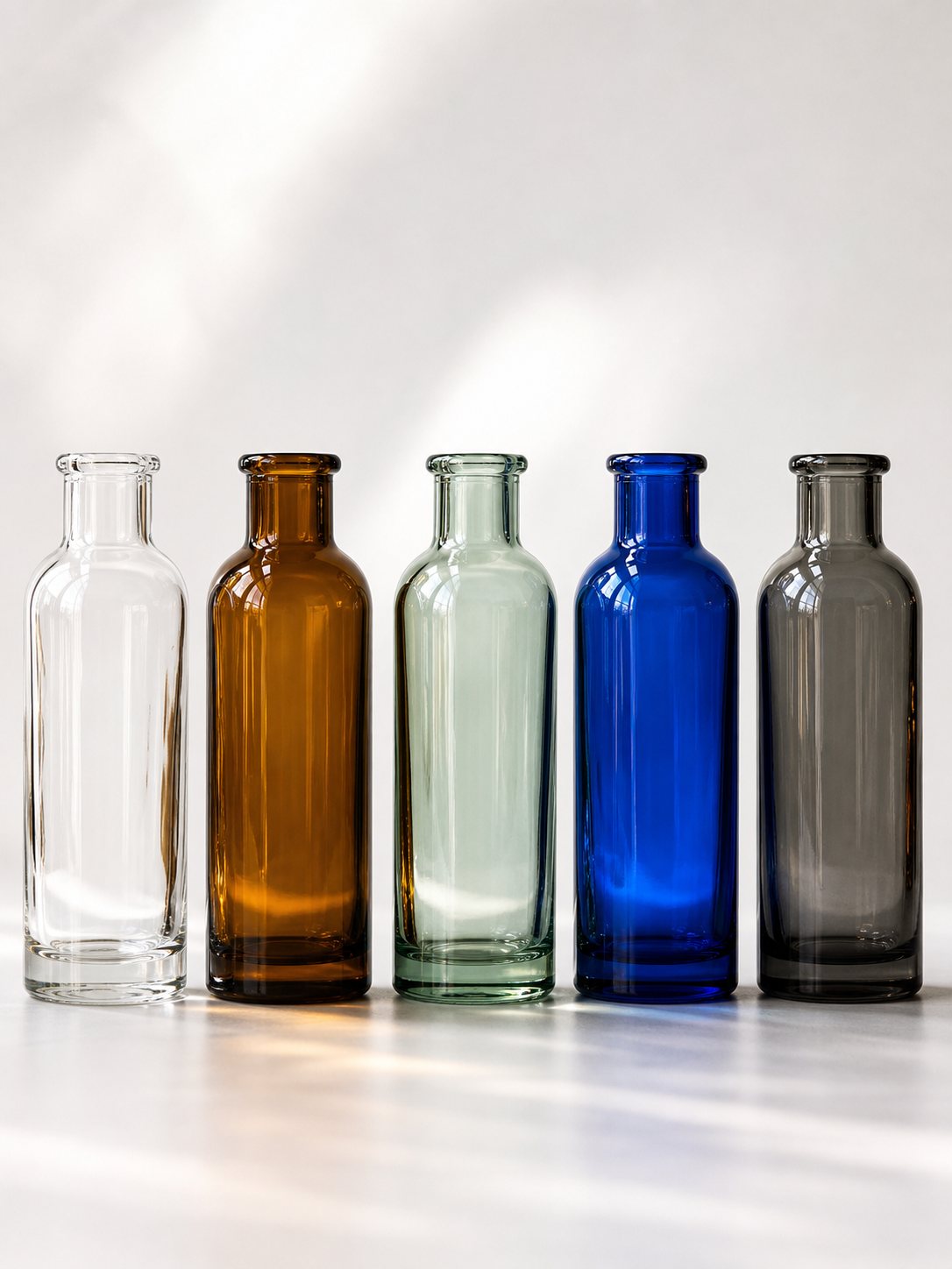 Five unlabeled glass bottles in clear, amber, pale green, cobalt blue, and smoked grey on a white surface.