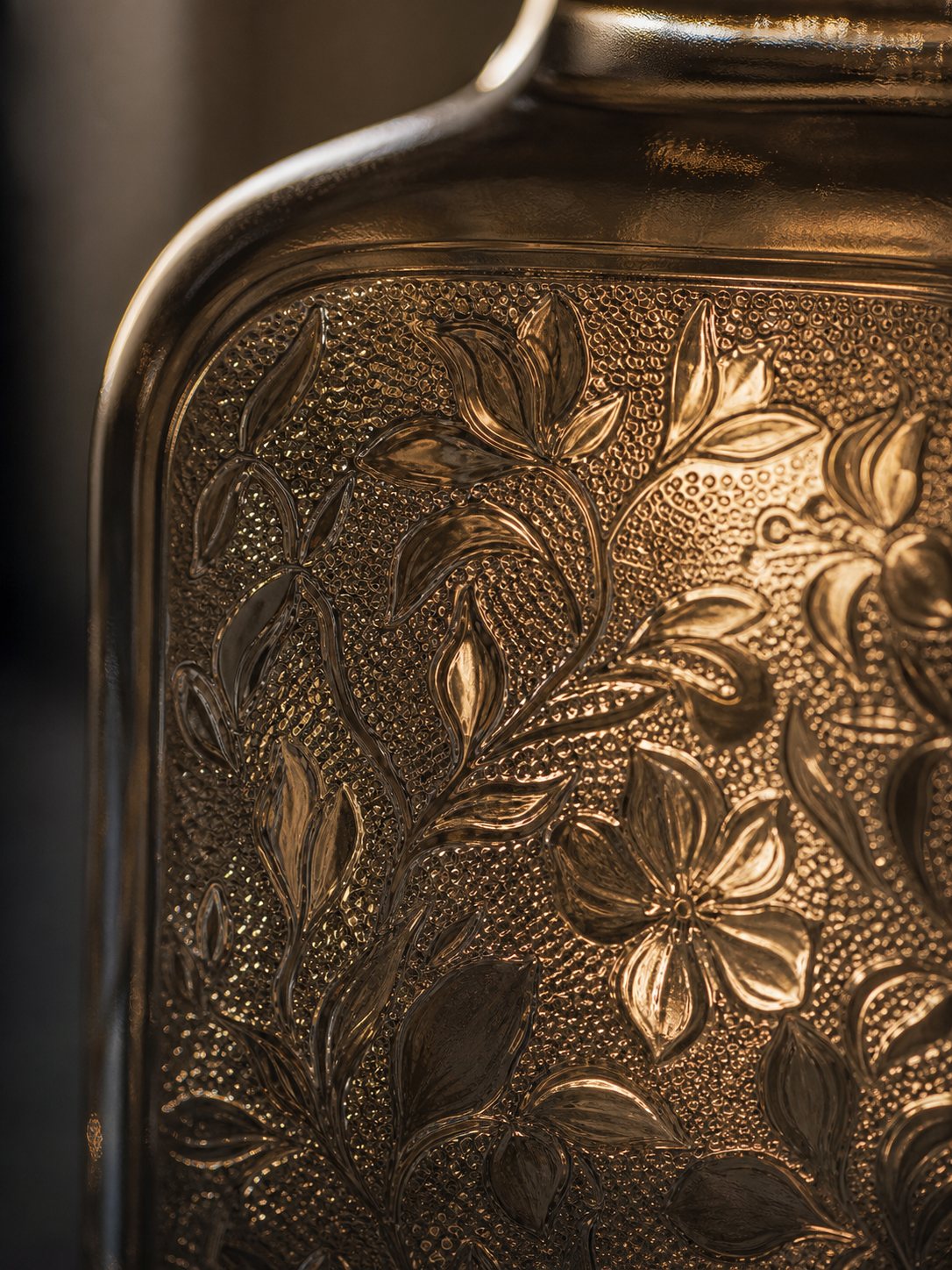 Close-up of a glass bottle surface with raised botanical texture and warm raking light.