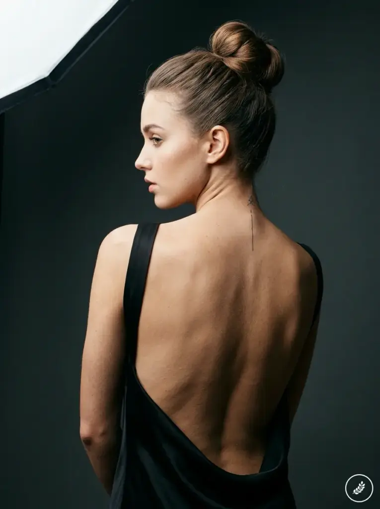 Woman_with_neck_tattoo_202607170037 Studio portrait of woman in backless black dress, hair in a bun, side profile showing bare back and small neck tattoo. | Sky Rye Design Studio portrait of woman in backless black dress, hair in a bun, side profile showing bare back and small neck tattoo.