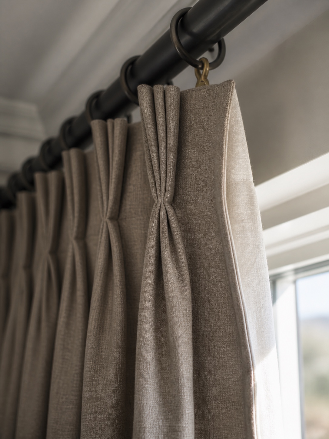 Pinch pleat drapery heading detail Close-up of a triple pinch pleat drapery heading with brass hardware and visible lining. | Sky Rye Design Close-up of a triple pinch pleat drapery heading with brass hardware and visible lining.
