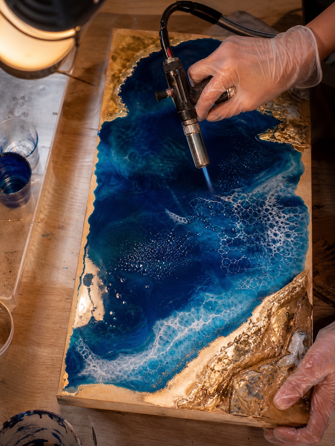 Gloved hands use a torch to remove bubbles from a blue and gold resin ocean pour.