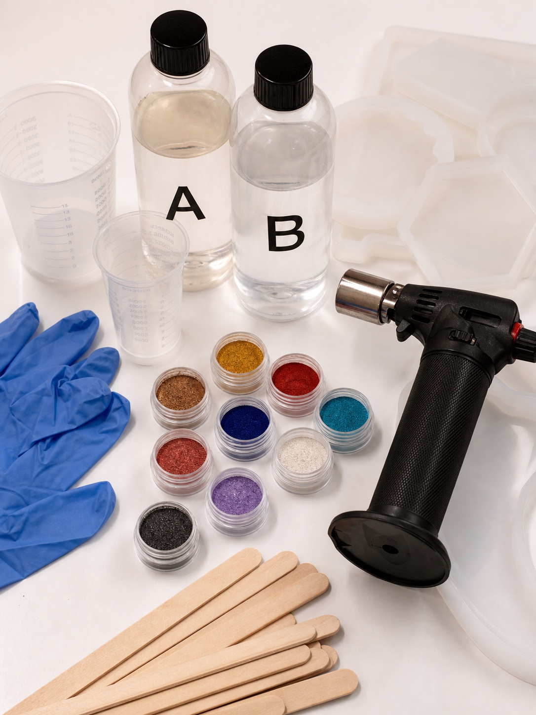 Flat lay of epoxy resin bottles, measuring cups, silicone molds, gloves, pigments and a torch.