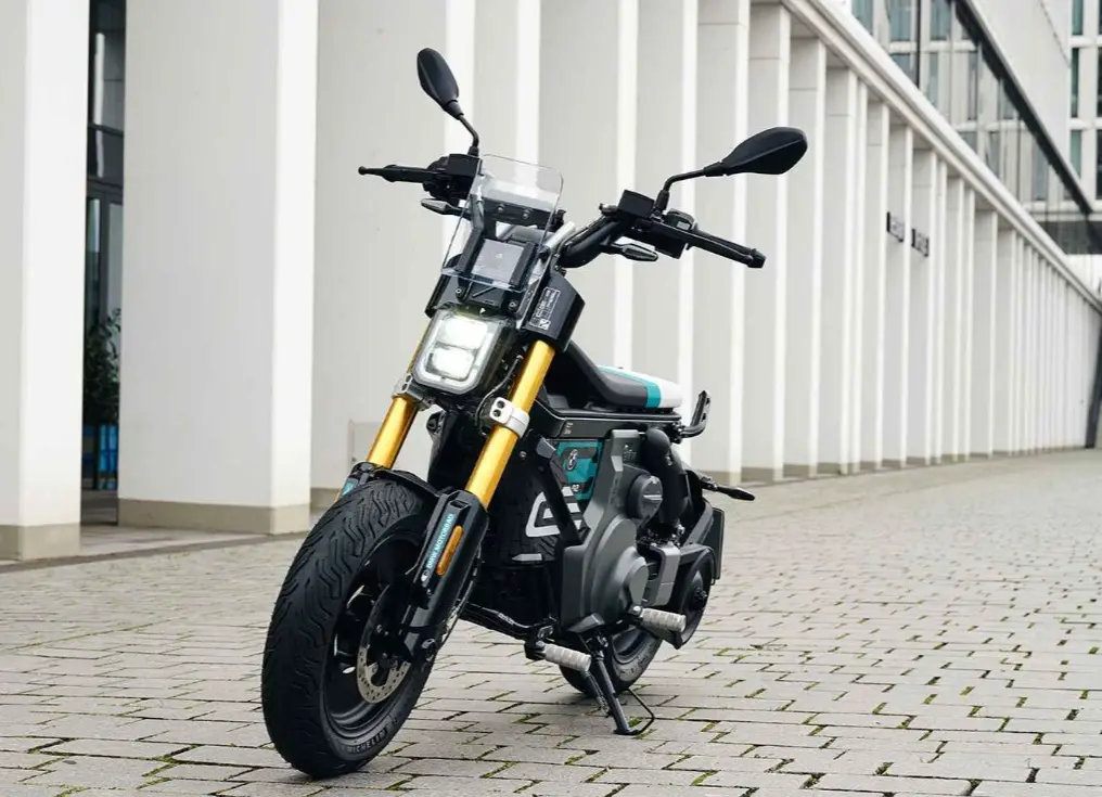 Black electric motorcycle with gold forks parked on paved plaza outside modern white-column building