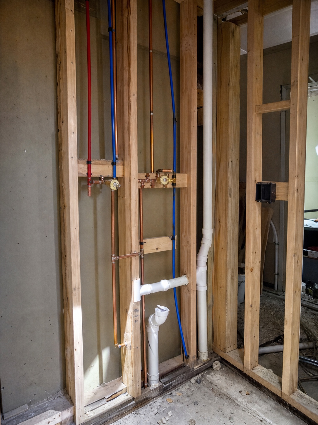 A bathroom wall at rough-in stage with exposed copper and PEX supply lines, drain pipes, and shut-off valves between studs.