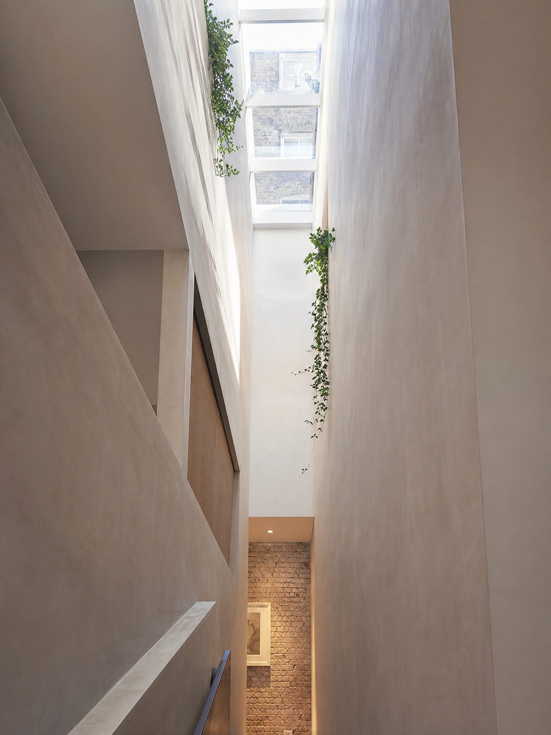 Central light well in a narrow urban home Bright vertical atrium with a skylight and hanging greenery along tall beige walls, ending at a brick accent wall and framed artwork. | Sky Rye Design Daylight pours through a central light well into a pale stairwell inside a narrow urban home.