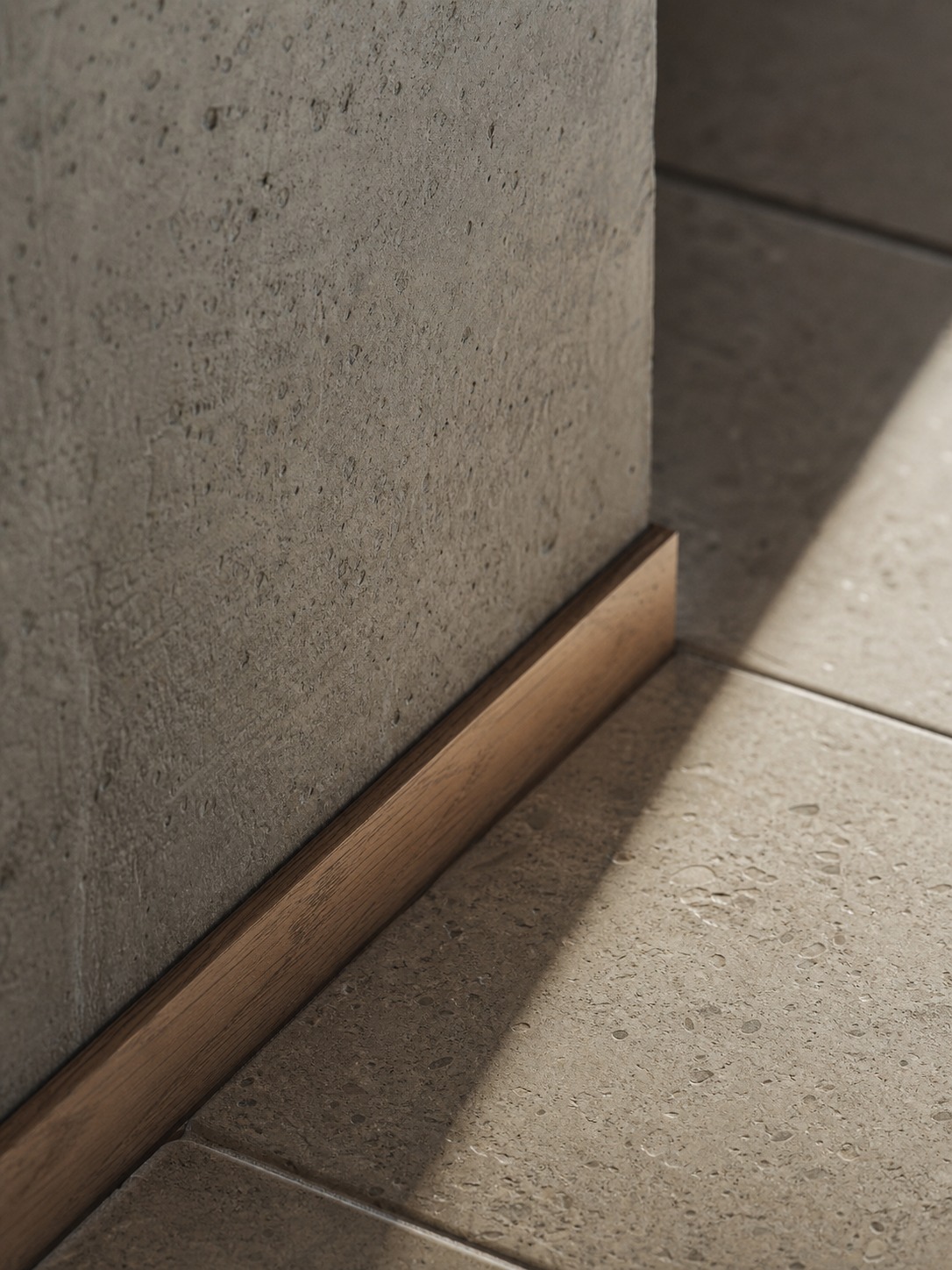 Close-up interior detail where concrete, timber skirting, and stone tile meet in raking side light.