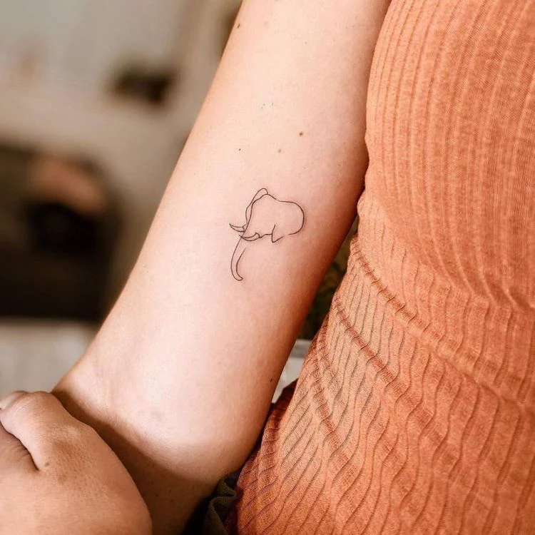 Small minimalist single-line elephant tattoo on woman's inner forearm, shown with ribbed orange tank top