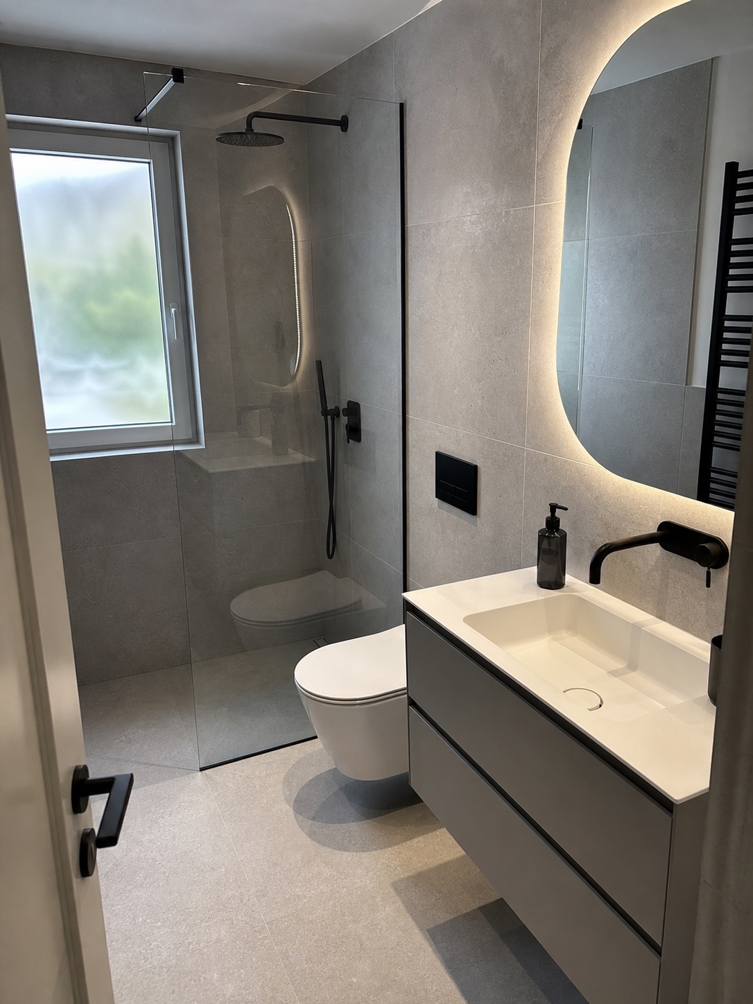 Small renovated bathroom with grey tile, wall-hung vanity, matte black fixtures, and a backlit mirror.