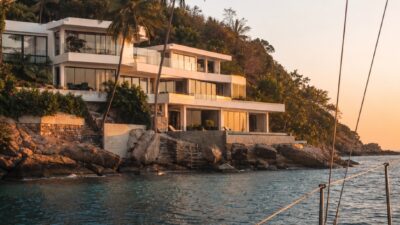 A modern tropical villa seen from offshore at golden hour, with rooflines, white concrete, glass, and coastline in view.