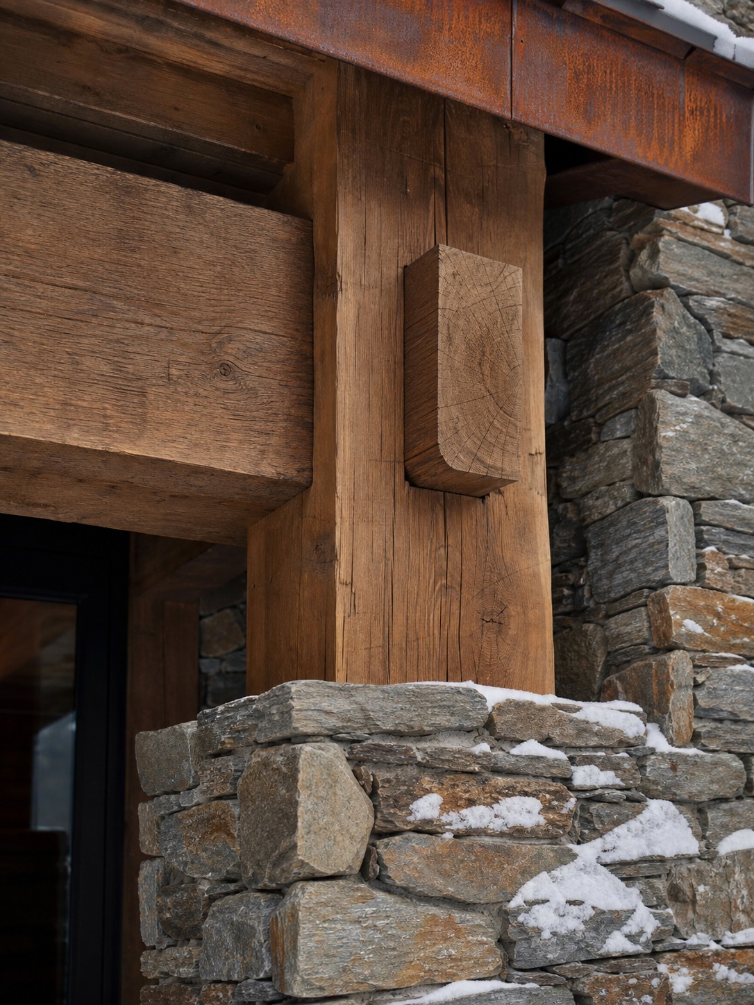 Close-up of a rough-sawn timber beam, stacked stone column, and weathering steel fascia with snow on the stone