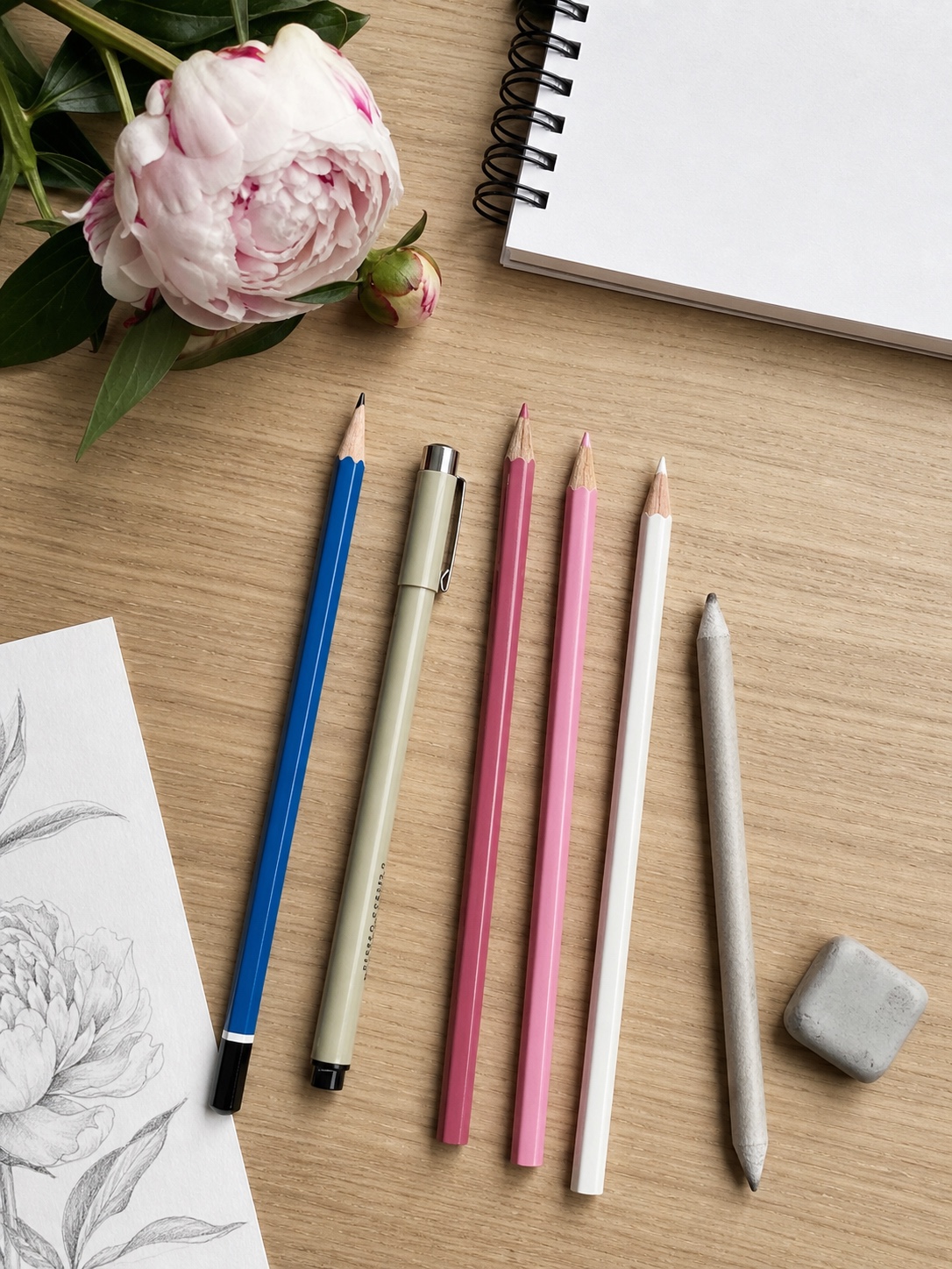 Botanical drawing materials arranged on a light desk with pencils, pen, eraser, blending stump, sketchbook, and peony reference.