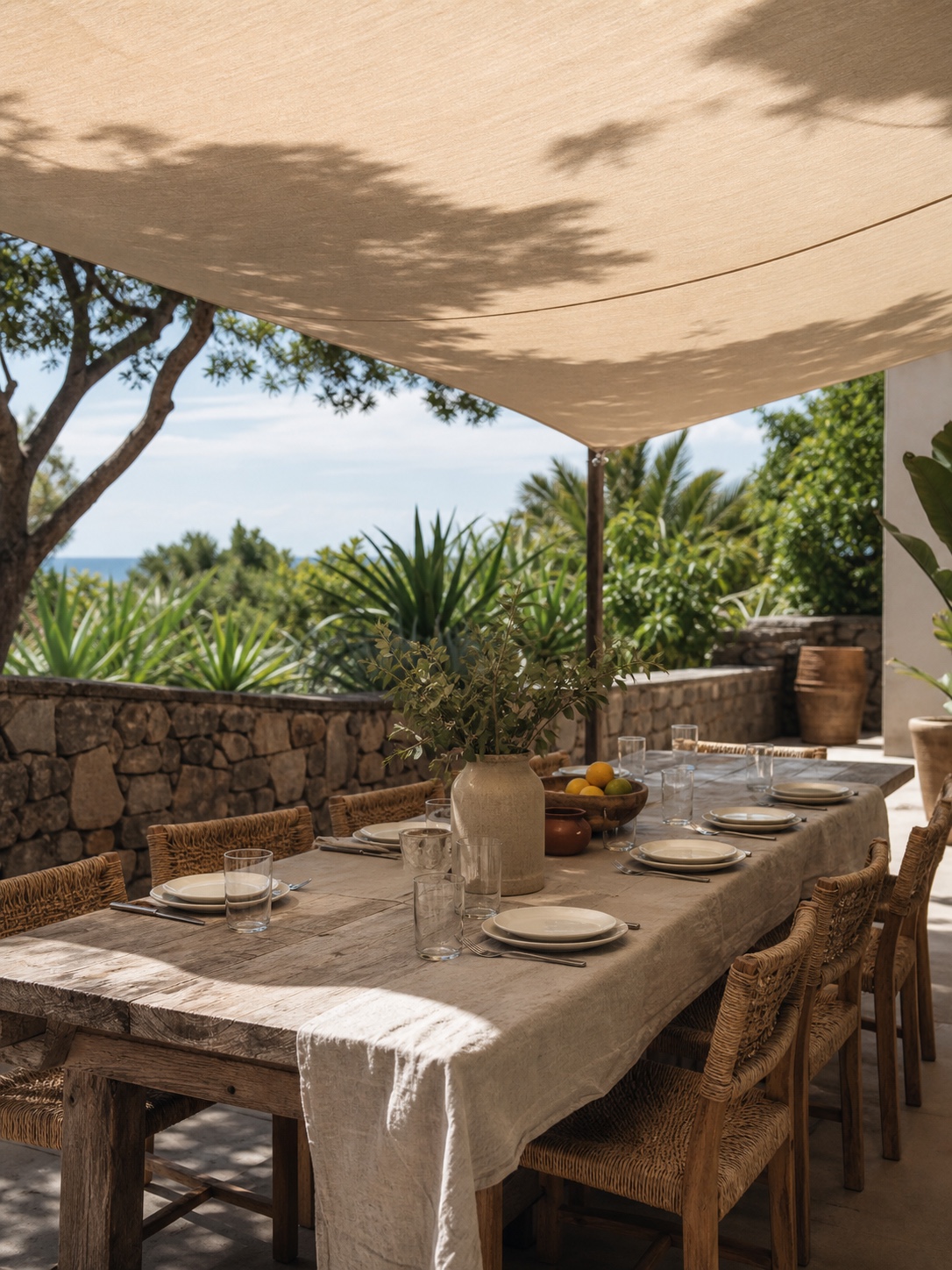 A shaded outdoor dining area under a canvas sail with a timber table and woven chairs.