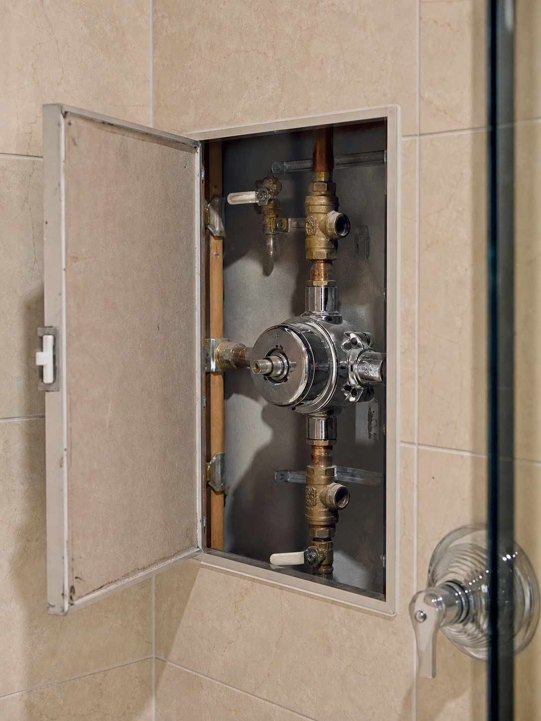 An open bathroom access panel exposing a chrome shower valve body, supply connections, and isolation valves behind tile.