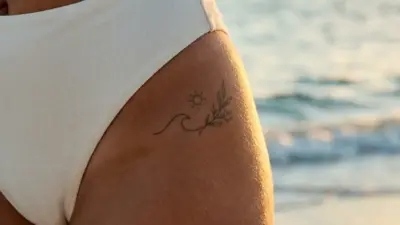Close-up of woman's hip in white bikini bottom with small sun, wave and leaf tattoo at beach sunset
