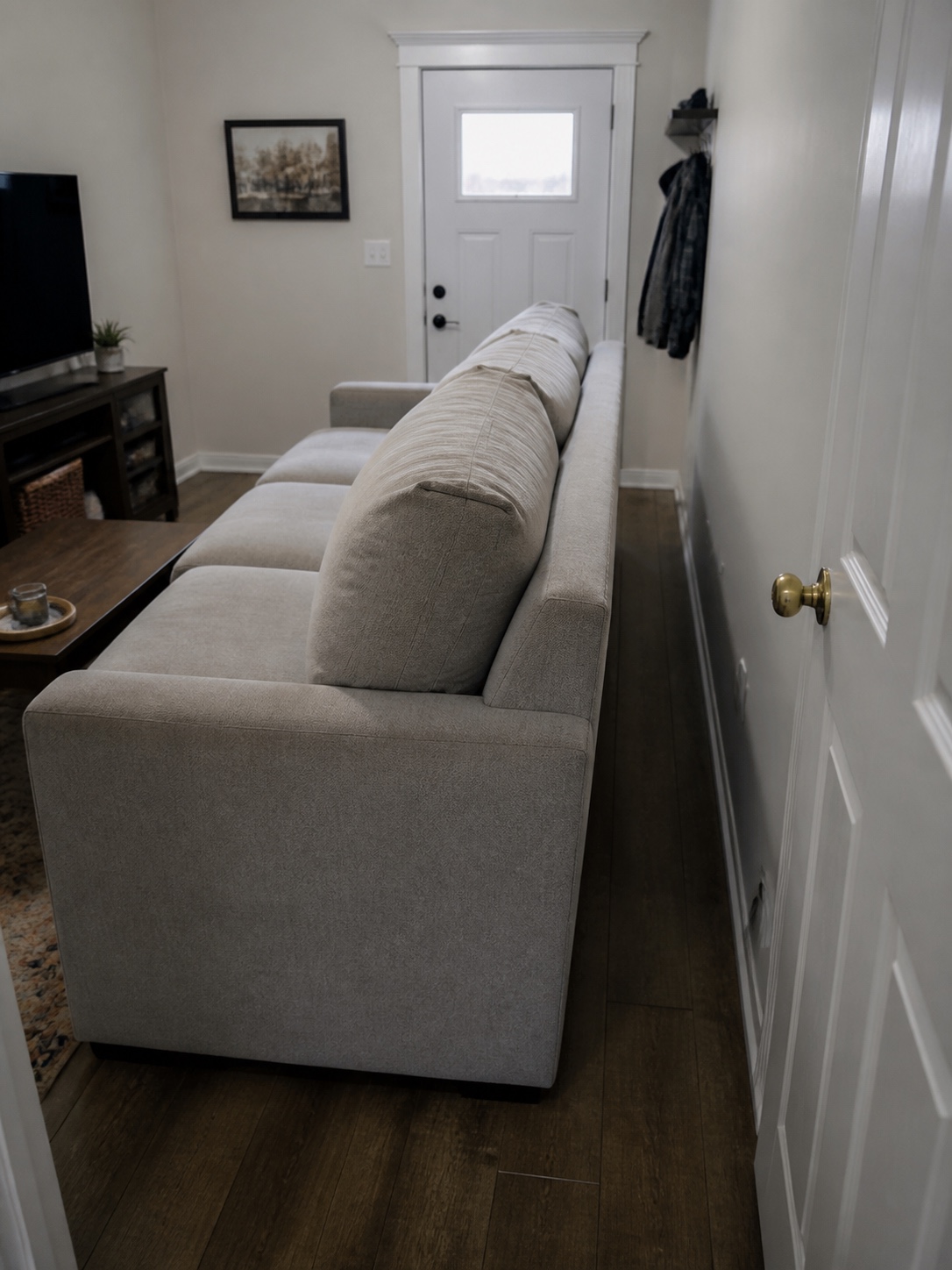 Sofa Too Large for Room Narrow living room with a long light-gray sofa along the wall, front door visible at the end, coat rack with a jacket to the right, and a TV setup on the left. | Sky Rye Design An oversized sofa blocks a doorway and leaves a narrow walkway in a small living room.