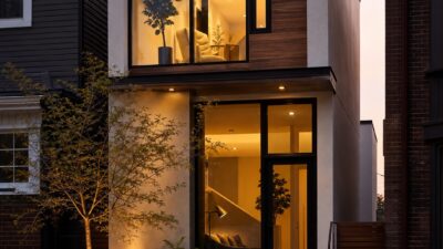 A compact two-story urban home glows warmly at dusk between close neighboring buildings.