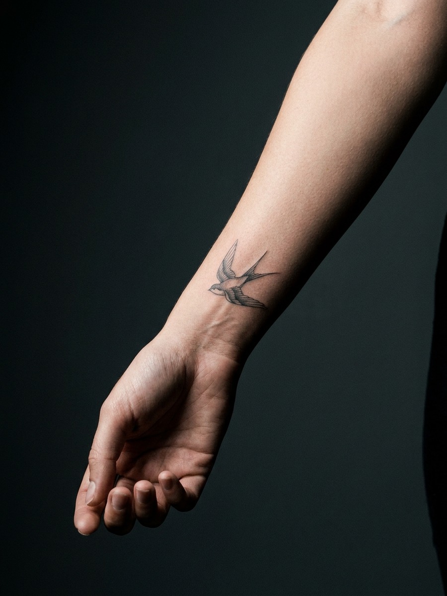Single-needle fine-line swallow tattoo mid-flight on a woman's forearm turned to present the inner wrist and forearm.