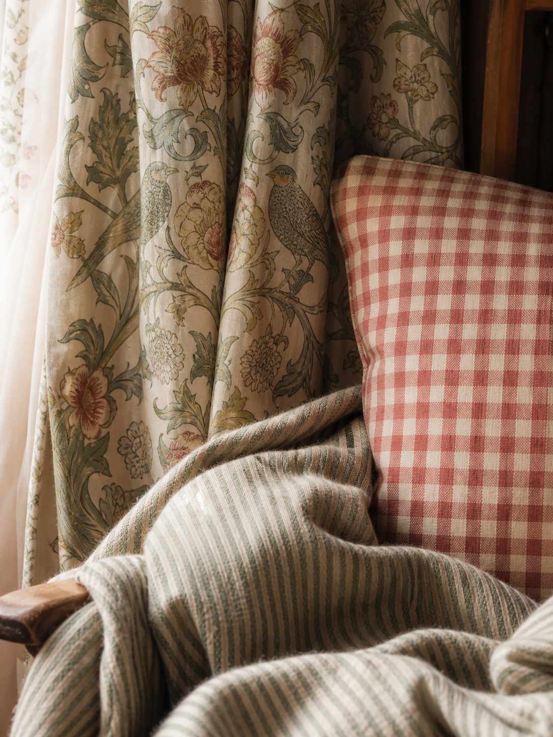 Layered English cottage textiles with floral curtain red gingham cushion ticking stripe blanket and muted sage palette Layered English cottage textiles with floral curtain, red gingham cushion, ticking stripe blanket, and muted sage palette. | Sky Rye Design Layered English cottage textiles with floral curtain, red gingham cushion, ticking stripe blanket, and muted sage palette.