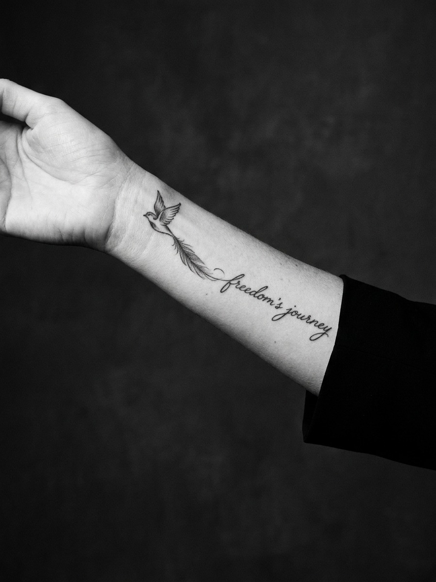 A small bird in flight with a single trailing feather that dissolves into fine cursive script along the forearm. on a woman's forearm.