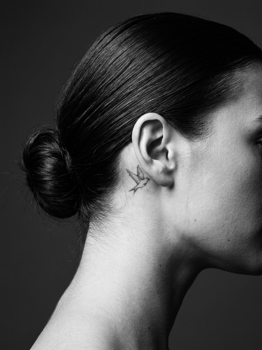 Close-up of a woman's neck and ear showing a small dove tattoo behind the ear.