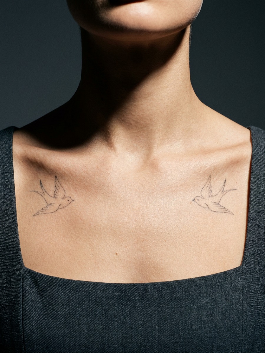Two mirrored small birds facing each other on a woman's collarbones and upper chest.