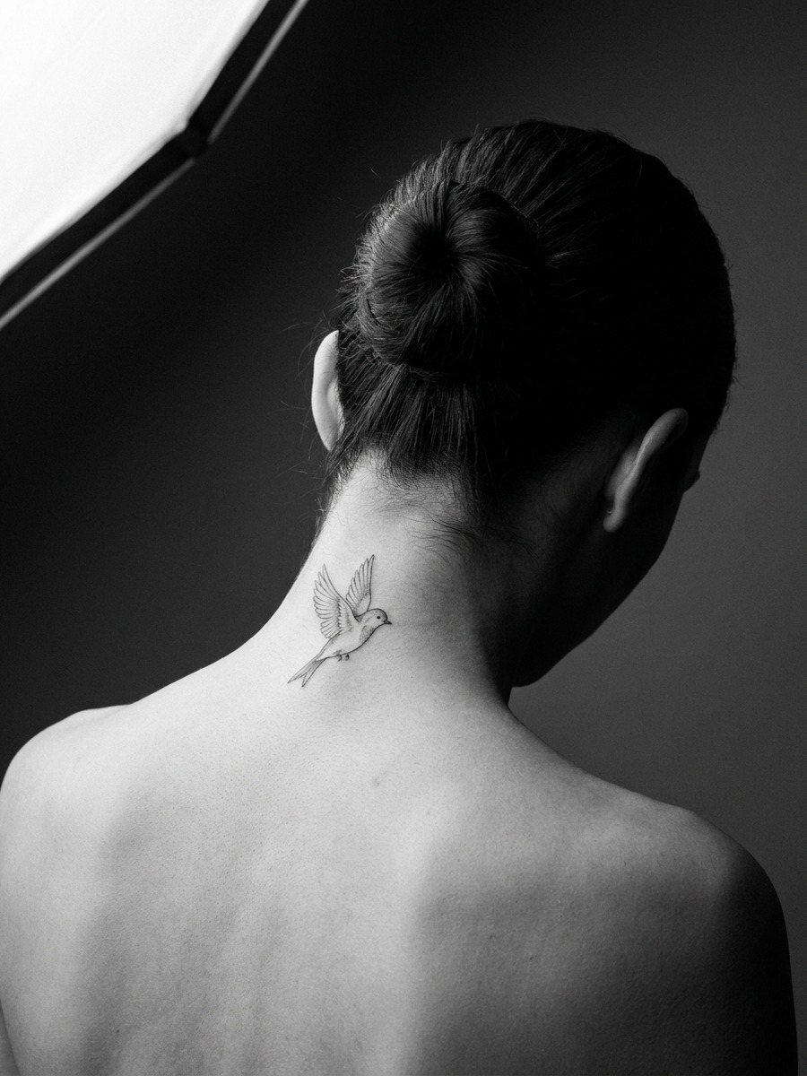 A small bird tattoo centered on the nape on a woman's nape and upper neck.