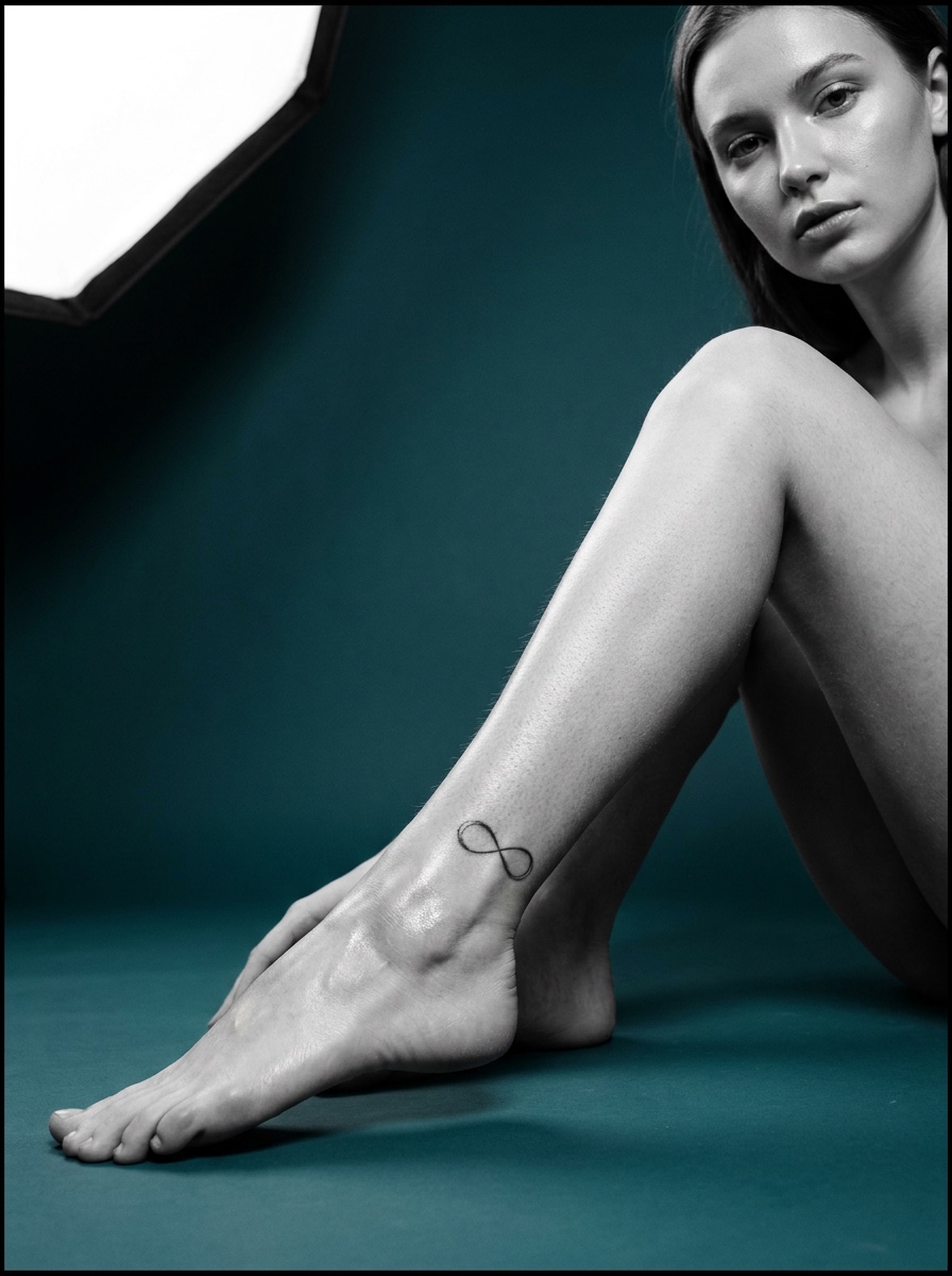 A small fine-line infinity tattoo on an arched ankle in teal studio light