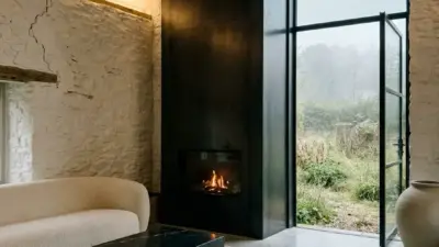 Cozy living room with a black fireplace, exposed wooden beams, and a large glass door opening to a garden, beige curved sofa, and a black marble coffee table on a woven rug.
