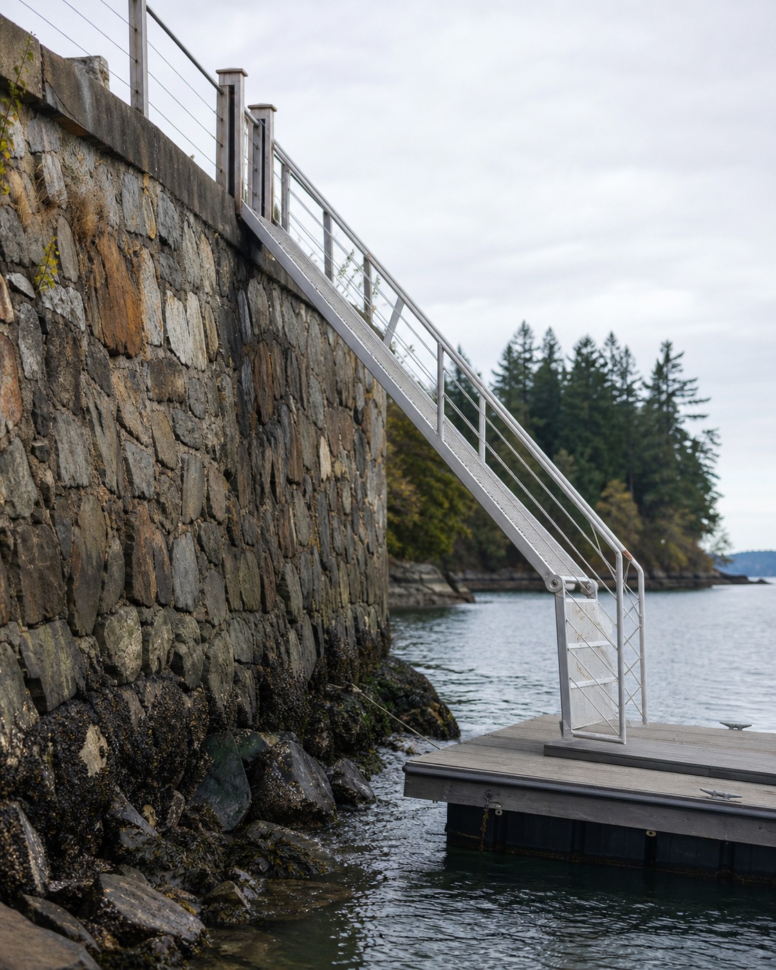 Hinged aluminum gangway descending from a stone retaining wall to a floating platform.