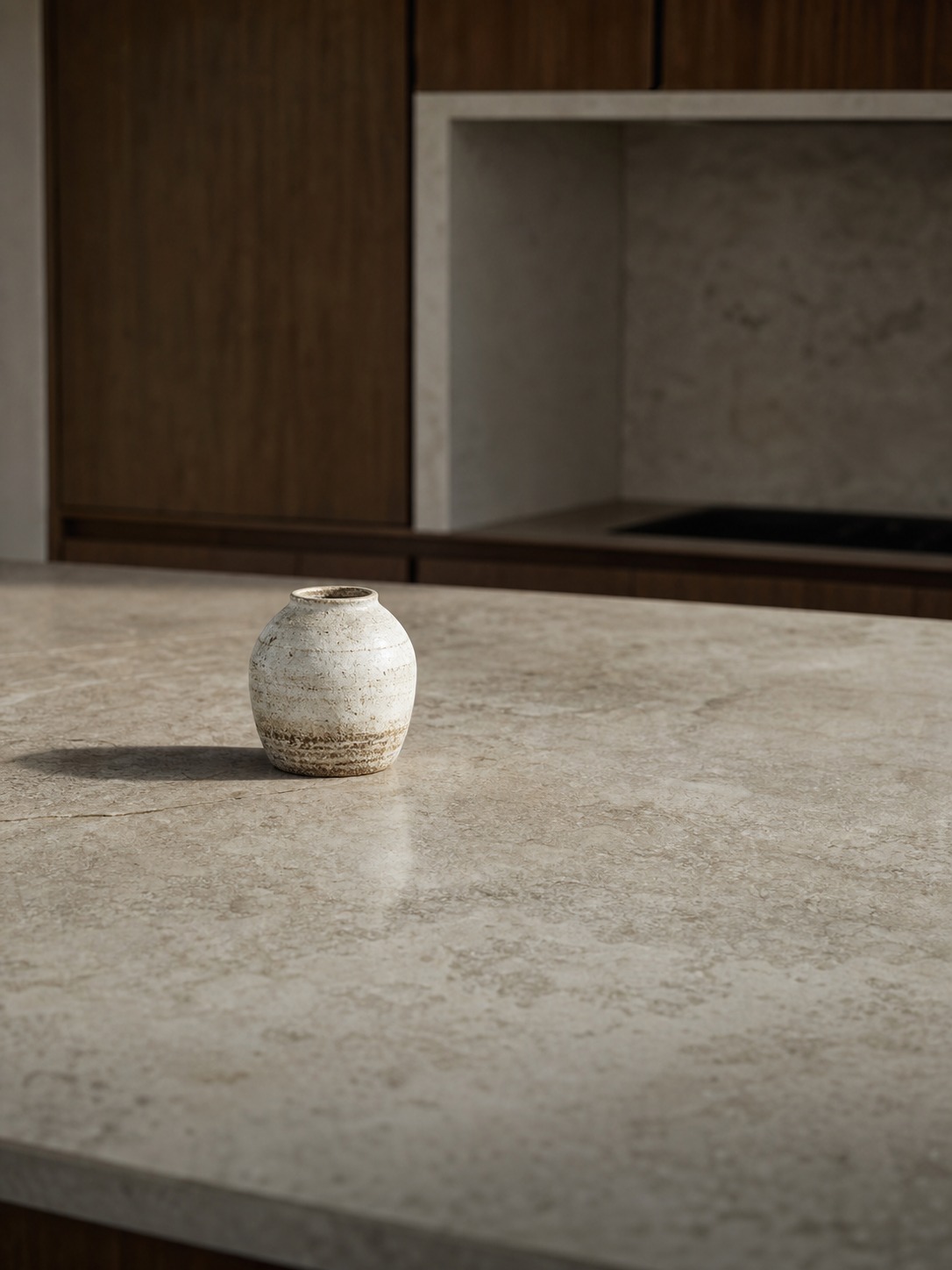 Handcrafted vessel on a stone kitchen island A small, speckled beige ceramic vase sits on a pale stone kitchen island with dark wood cabinets in the background. | Sky Rye Design Single handcrafted ceramic or stone vessel placed on an expansive stone kitchen island.