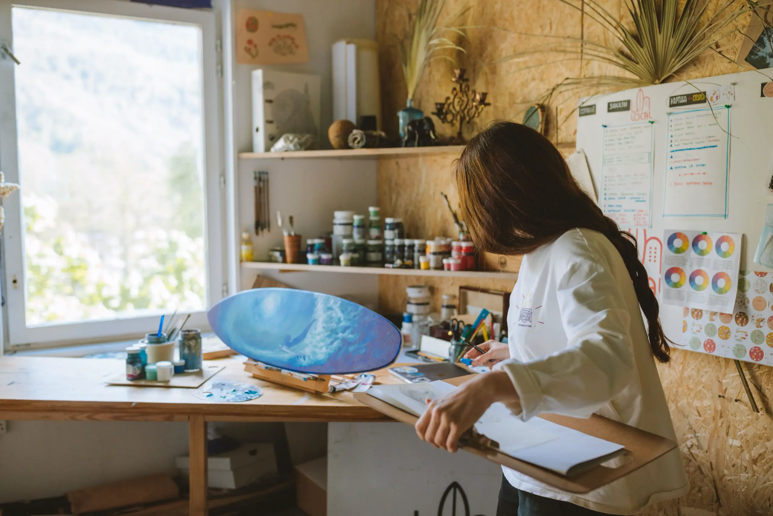 13f0ebec-49c4-4548-90ba-eaa1b5e4f57e Female artist painting a blue abstract canvas in a sunlit art studio with jars of paint and color wheel charts | Sky Rye Design Female artist painting a blue abstract canvas in a sunlit art studio with jars of paint and color wheel charts