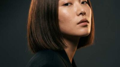 Editorial portrait of a model with a sharp blunt Korean bob haircut and glass-skin makeup against a charcoal grey studio backdrop