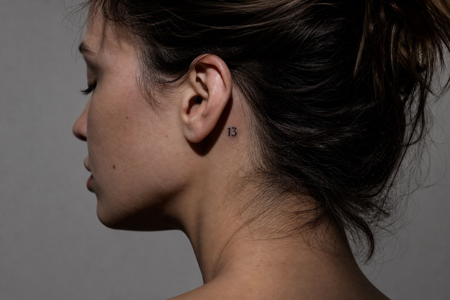 Minimal feminine behind-ear tattoo with the number 13 in fine black ink