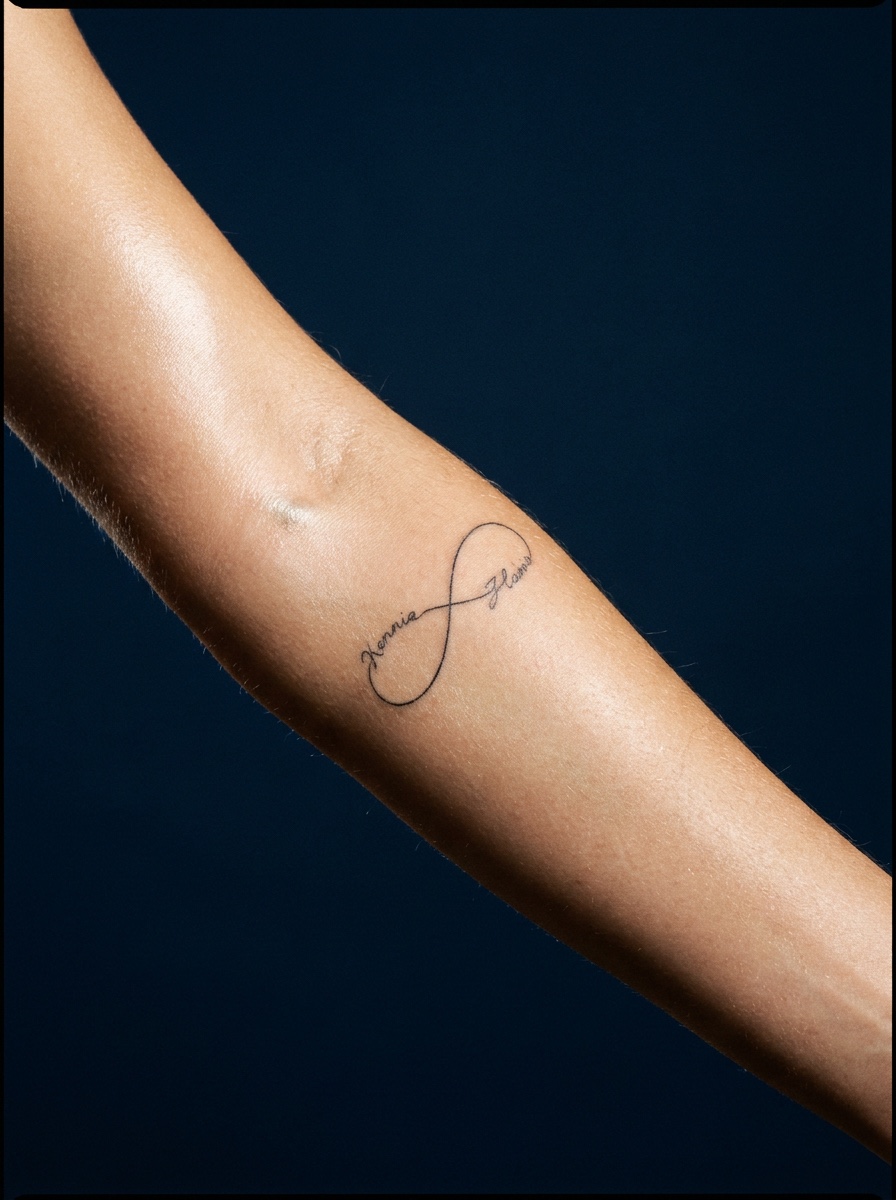 A fine-line infinity tattoo on the forearm with two names following the loops in script